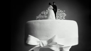 Figurines of a woman in a wedding dress and a man wearing a suit stand atop a wedding cake with their backs turned toward the camera.