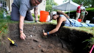 UMass Boston graduate students Nicholas Densley, of Missoula, Mont., left, and Kiara Montes, of Boston, right, use brushes while searching for artifacts at an excavation site, in this Wednesday, June 9, 2021 file photo, on Cole's Hill, in Plymouth, Mass.