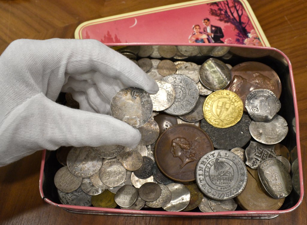 Image of the tin box where a rare coin from colonial Boston was found. A gloved hand holds the coin above the box.