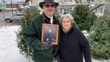 An elderly couple poses in front of Christmas trees, holding a picture of their late son