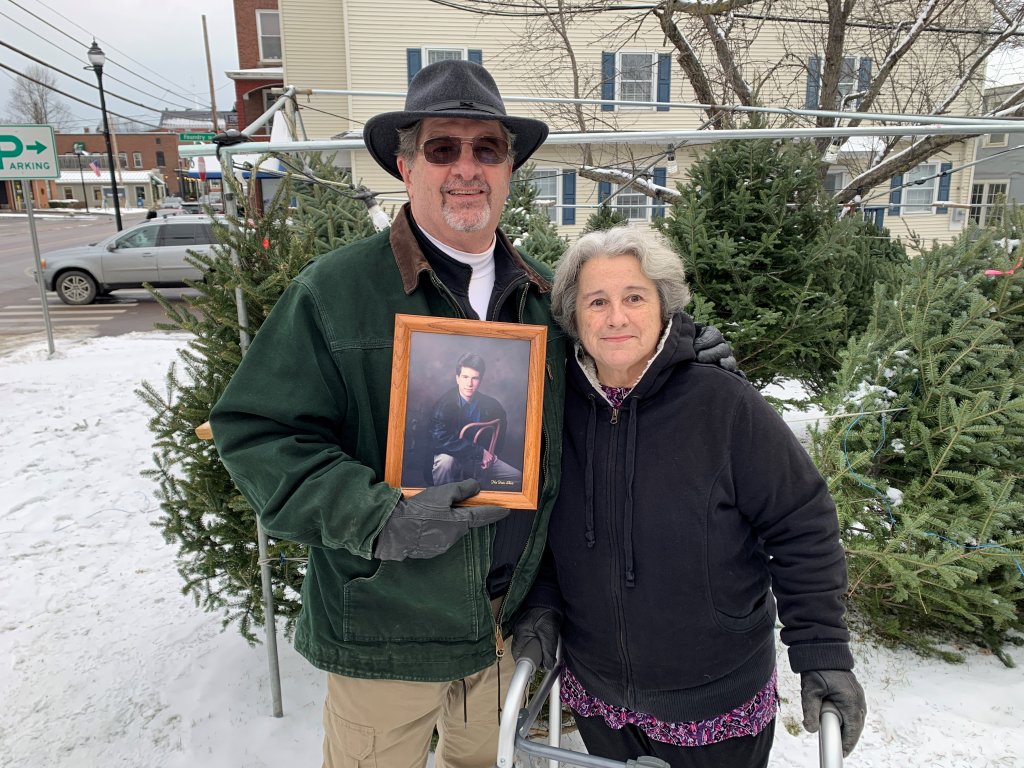 An elderly couple poses in front of Christmas trees, holding a picture of their late son 