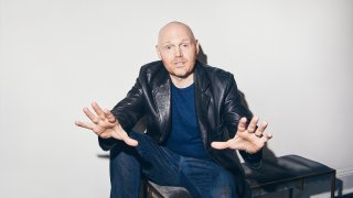 Comedian Bill Burr sits on a bench with his arms extended and hands open for the camera
