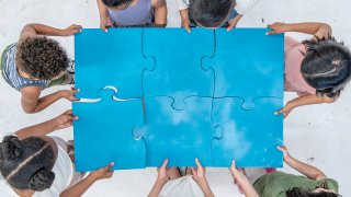 A large group of elementary students are seen each holding a puzzle piece as they fit them together with teamwork. They are each dressed casually as they focus on the task in this aerial view photo.