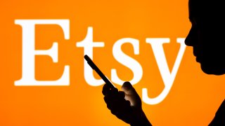In this photo illustration, the Etsy logo is seen in the background of a silhouetted woman holding a mobile phone.