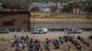 FILE - Migrants sit in rows in front of U.S. security forces in the area between the two walls that separate Mexico from the United States.