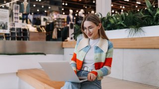 A young female freelancer influencer in glasses and fashionable casual clothes works online remotely using a laptop while sitting on the stairs in a shopping mall in a public place.