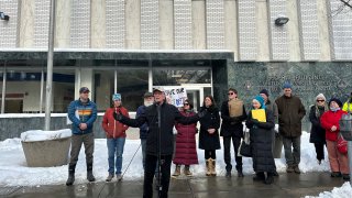 A man (Sen. Peter Welch) speaks in front of a crowd of people, who have gathered in front of a post office. Snow is on the ground.
