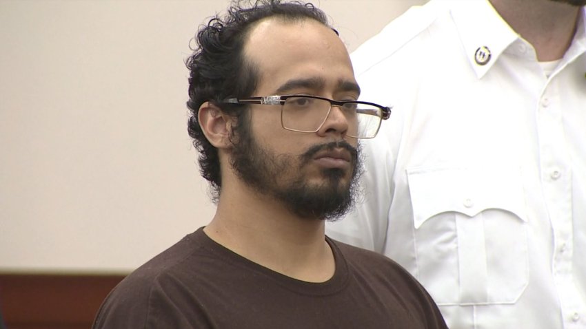 A man stands in a courtroom. He has dark curly hair,a beard and wears glasses and a brown t-shirt