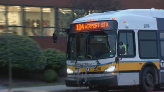 A white and yellow MBTA bus drives down the street. The screen at the top of the windshield reads "104 Airport Sta"